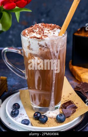 Lait d'amande chocolat chaud recouvert de crème fouettée dans un verre latte, bleuets et morceaux de chocolat autour de lui, fleurs derrière lui. Banque D'Images