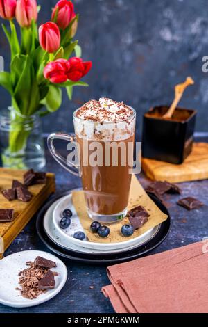 Chocolat chaud recouvert de crème fouettée dans un verre latte, bleuets et morceaux de chocolat autour de lui, fleurs derrière lui. Banque D'Images