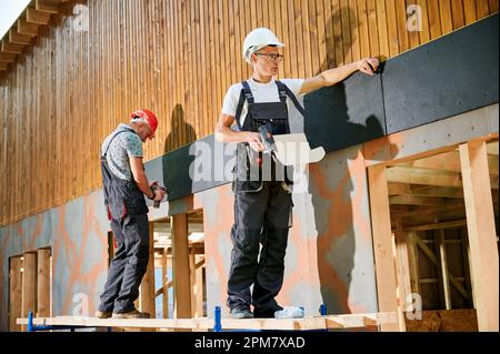 Charpentiers construisant une maison encadrée en bois. Deux hommes ouvriers claviant la façade de la maison avec des panneaux de particules de ciment, les fixant avec des tournevis. Concept de construction écologique moderne. Banque D'Images