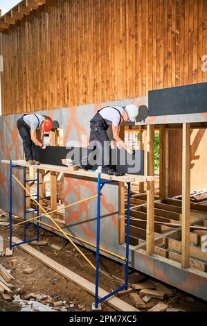 Charpentiers construisant une maison encadrée en bois. Deux hommes ouvriers claviant la façade de la maison avec des panneaux de particules de ciment, les fixant avec des tournevis. Concept de construction écologique moderne. Banque D'Images