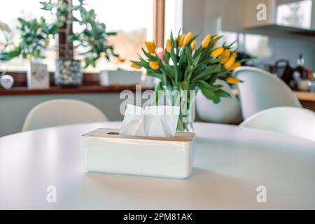 Boîte de mouchoirs sur la table du salon. Fleurs printanières dans un vase en verre sur une table en bois blanc dans la salle de séjour. Bouquet de tulipes jaunes. Banque D'Images