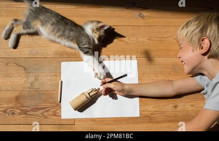 joyeux enfant tire avec des crayons couchés sur le sol dans une chambre lors d'une journée ensoleillée en compagnie d'un chaton bien-aimé. Amitié et jeu d'un enfant Banque D'Images
