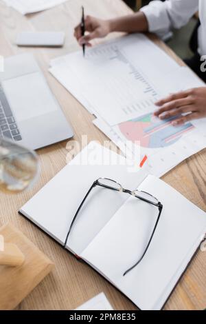 vue à grand angle des lunettes et carnet vierge près d'une femme d'affaires afro-américaine rognée travaillant avec des infographies au bureau, image de stock Banque D'Images