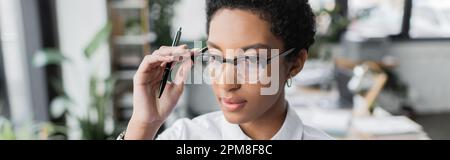 jeune femme d'affaires afro-américaine tenant un stylo et ajustant des lunettes de vue stylées dans le bureau, bannière, image de stock Banque D'Images