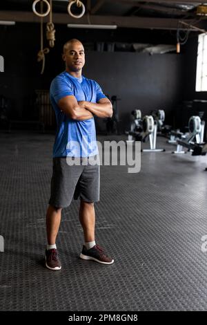 Jeune homme plein de confiance biraciale avec tête rasée croisant les bras et debout dans la salle de gym Banque D'Images