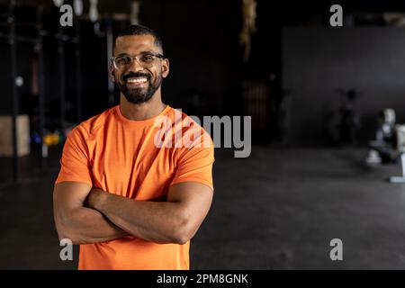 Portrait du jeune homme biracial barbu avec les bras croisés souriant et debout dans la salle de gym Banque D'Images