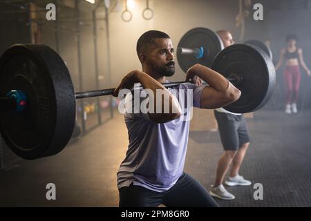 Jeune homme barbu biracial dédié levant la barbell en étant debout dans la salle de gym Banque D'Images