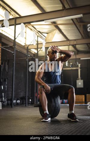 Pleine longueur de jeune biracial fatigué essuyant le front tout en étant assis sur la barbell dans la salle de gym, l'espace de copie Banque D'Images