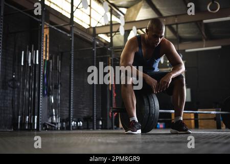 Pleine longueur de jeune biracial fatigué avec la tête rasée assis sur la barbell dans la salle de gym, espace de copie Banque D'Images