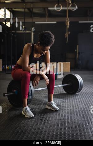 Pleine longueur de jeune femme biracial fatigué avec cheveux afro assis sur la barbell dans la salle de gym, espace copie Banque D'Images