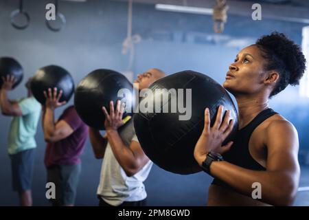 Jeune femme et homme biracial s'exerçant avec un ballon de fitness noir tout en se tenant debout dans une rangée à la salle de gym Banque D'Images