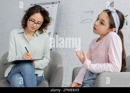 Psychologue écrivant sur le presse-papiers tout en brouillé enfant parlant dans la salle de consultation, image de stock Banque D'Images