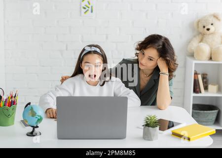Préteen fille collant hors langue pendant la leçon en ligne de thérapie de la parole sur ordinateur portable près de la mère à la maison, image de stock Banque D'Images
