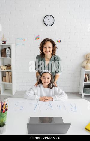 Mère souriante et fille de la préadolescence regardant l'appareil photo près des papiers d'ordinateur portable avec des lettres sur le bureau, image de stock Banque D'Images