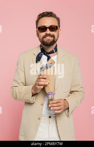 homme plein de joie dans des lunettes de soleil stylées et foulard de cou tenant des fleurs dans papier d'emballage isolé sur rose, image de stock Banque D'Images