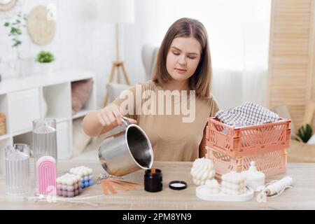 femme faisant des bougies à la maison, versant de la cire liquide jaune de la casserole dans la forme de bouteille de verre foncé Banque D'Images