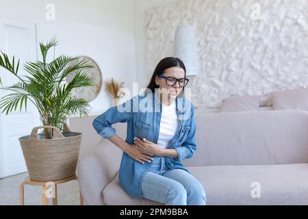 Une jeune femme est assise sur le canapé à la maison, grimaquant et tenant son estomac. Souffre de douleurs abdominales sévères, de douleurs menstruelles, de problèmes de grossesse, de fausse couche, d'empoisonnement, spasmes. Banque D'Images