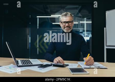 Portrait d'un vieil homme aux cheveux gris, d'un comptable, d'un auditeur assis au bureau à un ordinateur portable et d'une calculatrice. Il travaille avec des documents, vérifie des comptes. Sourire à l'appareil photo. Banque D'Images