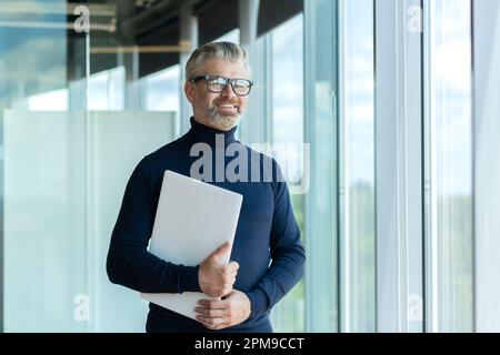 Portrait d'un ancien concepteur, architecte, ingénieur avec une vaste expérience de travail. Il se tient dans le bureau près de la fenêtre avec un ordinateur portable dans ses mains. Regardez l'appareil photo avec assurance et sourire. Banque D'Images