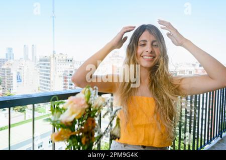 portrait d'une jeune femme latine d'origine colombienne, vêtue de jaune, assise sur son balcon d'appartement, confondue avec les mains sur la tête, souriant et lo Banque D'Images