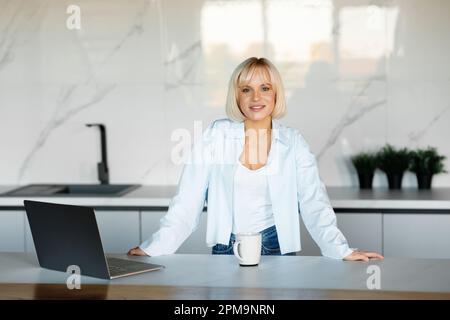 Une jeune fille blonde travaille sur un ordinateur portable à la maison dans la cuisine. Il y a un éclairage naturel, elle porte un T-shirt blanc et une chemise bleue. Banque D'Images