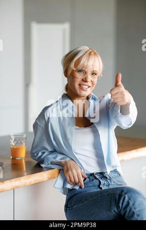 Une jeune fille blonde dans des verres élégants se trouve dans la cuisine avec un verre de jus. Regarde l'appareil photo et montre le geste super. Banque D'Images