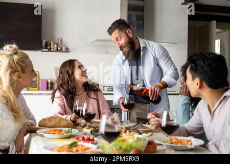 Les amis se rassemblent pour avoir de la nourriture italienne ensemble, un jeune homme barbu verse du vin rouge en verre à une jeune femme Banque D'Images