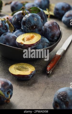 Mise au point sélective, faible profondeur de champ. Prunes mûres avec feuilles et moitiés de prunes dans un bol en céramique sur une table grise, à côté d'un couteau, sélec. Gros plan Banque D'Images