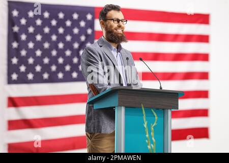 Homme barbu sur un podium en pronontant un discours et souriant avec le drapeau américain en arrière-plan Banque D'Images