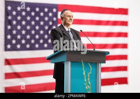 Homme politique américain prononcera un discours sur un podium en souriant avec un drapeau en arrière-plan Banque D'Images