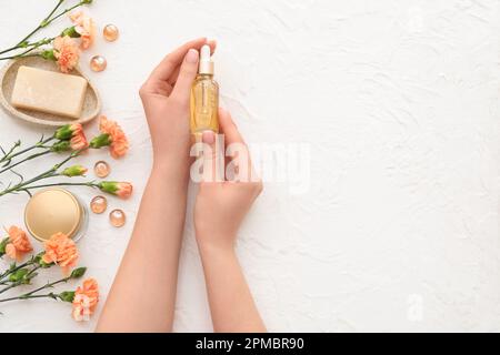 Mains de femmes avec une bouteille d'huile de cuticule, un produit cosmétique, du savon et des fleurs de carnation sur fond clair Banque D'Images