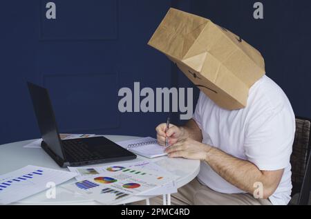 Un homme - un homme d'affaires dans une chemise blanche avec un sac de papier sur sa tête, avec un triste visage smiley dessiné, travaille à un ordinateur avec des documents, fait des notes dans Banque D'Images