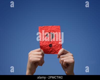 Un garçon tenant une note de papier rouge froissé avec un point d'interrogation dans ses mains contre le ciel bleu. Gros plan. Banque D'Images