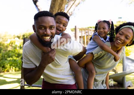 Parents afro-américains souriants qui coggysoutiennent leur fils et leur fille tout en se tenant debout dans l'aire de jeux Banque D'Images