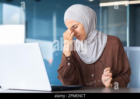Femme d'affaires fatiguée et surtravaillée dans le hijab sur le lieu de travail, femme musulmane massant les yeux avec des lunettes de vue, travailleuse travaillant tard sur le projet. Banque D'Images