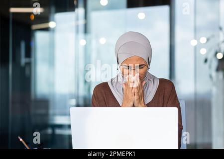 Femme d'affaires malade sur le lieu de travail, femme musulmane dans le hijab éternuant et ayant un nez qui coule, femme employée dans le bureau essuyant le nez avec des tissus, travaillant avec un ordinateur portable. Banque D'Images