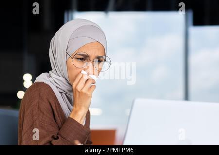 Femme d'affaires malade sur le lieu de travail, femme musulmane dans le hijab éternuant et ayant un nez qui coule, femme employée dans le bureau essuyant le nez avec des tissus, travaillant avec un ordinateur portable. Banque D'Images