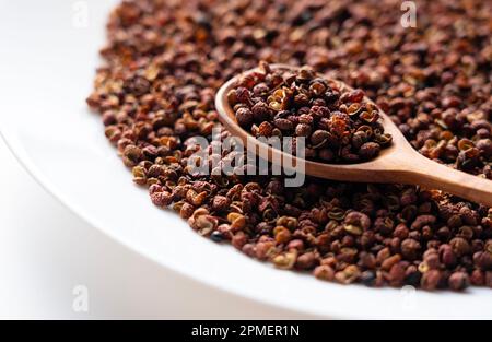 Poivre du Sichuan, cuillère en bois et assiette sur fond blanc. Le poivre du Sichuan est un membre de la famille des sansho utilisés dans la cuisine chinoise. Banque D'Images