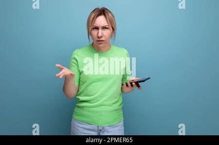 indignée belle fille blonde avec un téléphone dans sa main distribue ses mains sur un fond bleu avec un espace de copie Banque D'Images