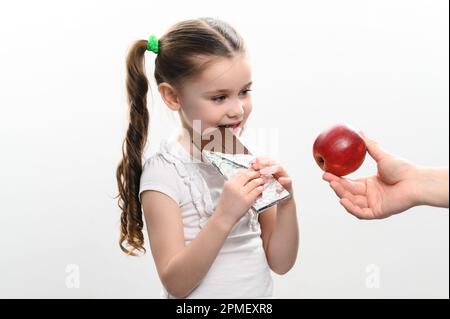 Portrait d'une petite fille sur fond blanc, elle choisit le chocolat au lieu d'une pomme. Banque D'Images