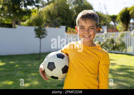 Portrait d'un adorable garçon caucasien souriant et tenant le ballon de football tout en se tenant sur une terre herbeuse dans la cour Banque D'Images