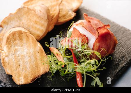 Fromage Camembert cuit au bacon. Morceaux croustillants de baguette et tranches de piment sur un panneau en pierre noire Banque D'Images