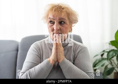 Frowning bouleversé femme mature mettre la tête sur les mains pliées, en regardant dans la distance. Perdu dans les pensées bouleverser la femme assise seule, tête tourné gros plan portrait Banque D'Images