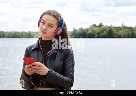 La jeune femme de race blanche, dans un casque bleu rouge, écoute de la musique et tape sur le smartphone sur la rive de la rivière. Portrait photo de Happy hippster fille rel Banque D'Images