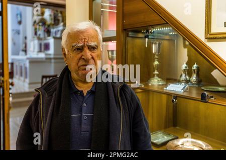 Un homme âgé se tient dans une pièce attenante à l'église Saint-Georges à Kormakitis (Korucam), République turque de Chypre du Nord (RTNC), devant une vitrine contenant de l'argenterie liturgique, dont un calice. L'intérieur de l'église est visible à travers une porte ouverte en arrière-plan. Banque D'Images