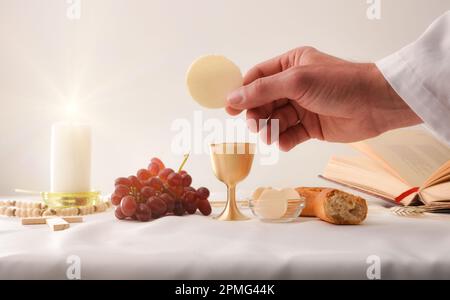 Mains d'un prêtre consacrant une armée comme corps du Christ pour la distribuer aux communiants avec table et objets sacrés derrière. Banque D'Images