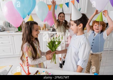 joyeux garçon donnant des tulipes à joyeux anniversaire fille près des amis sur fond flou, image de stock Banque D'Images