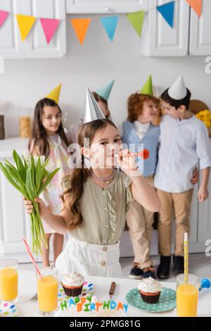 fille gaie tenant des tulipes et la corne de fête de soufflage près des amis pendant la fête d'anniversaire à la maison, image de stock Banque D'Images