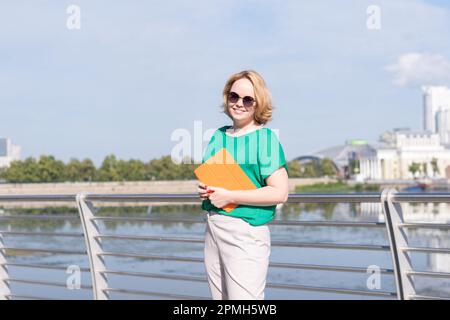 Une jeune fille souriante de blogueur en lunettes de soleil tenant un ordinateur portable ou une tablette dans ses mains sur le remblai sur le remblai. Une jeune fille marche dans la ville Banque D'Images