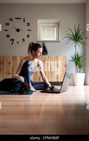 Jeune femme vêtue de vêtements décontractés, assise sur un tapis dans la salle de séjour à l'aide d'un ordinateur portable. Son chien noir allongé à côté d'elle. Banque D'Images